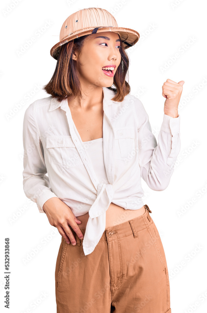 Young beautiful chinese girl wearing explorer hat smiling with happy face looking and pointing to the side with thumb up. Young beautiful chinese girl wearing explorer hat smiling with happy face looking and pointing to the side with thumb up.