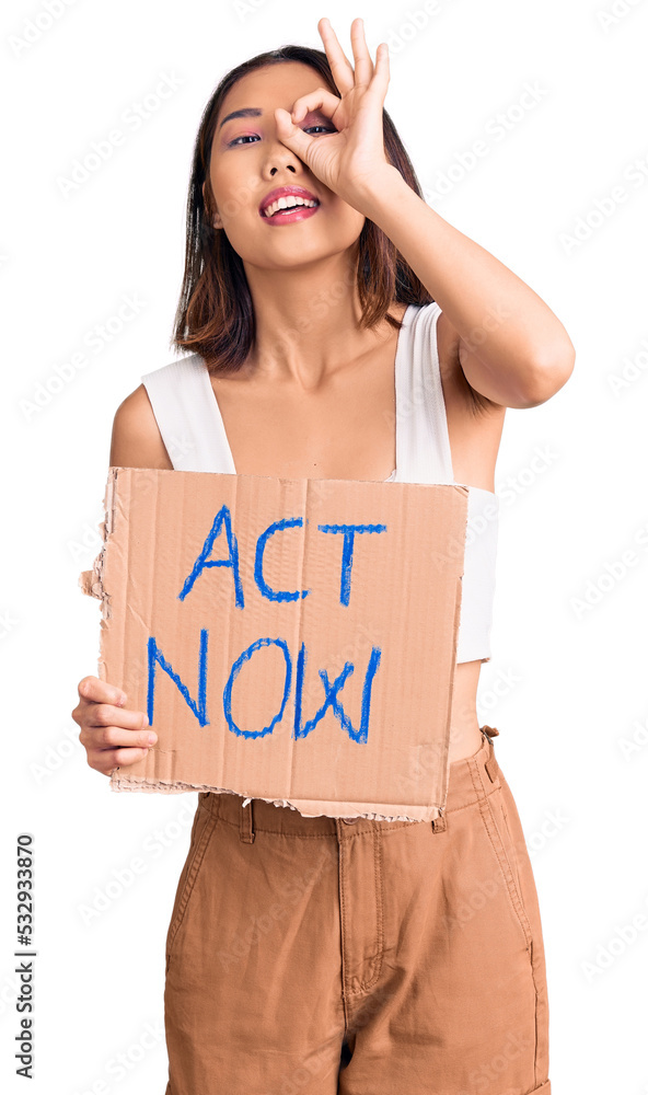 Young beautiful chinese girl holding act now banner smiling happy doing ok sign with hand on eye looking through fingers Young beautiful chinese girl holding act now banner smiling happy doing ok sign with hand on eye looking through fingers