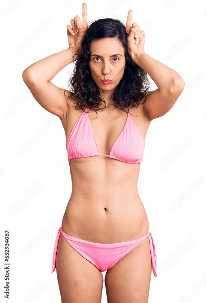 Young beautiful hispanic woman wearing bikini doing funny gesture with finger over head as bull horns Young beautiful hispanic woman wearing bikini doing funny gesture with finger over head as bull horns