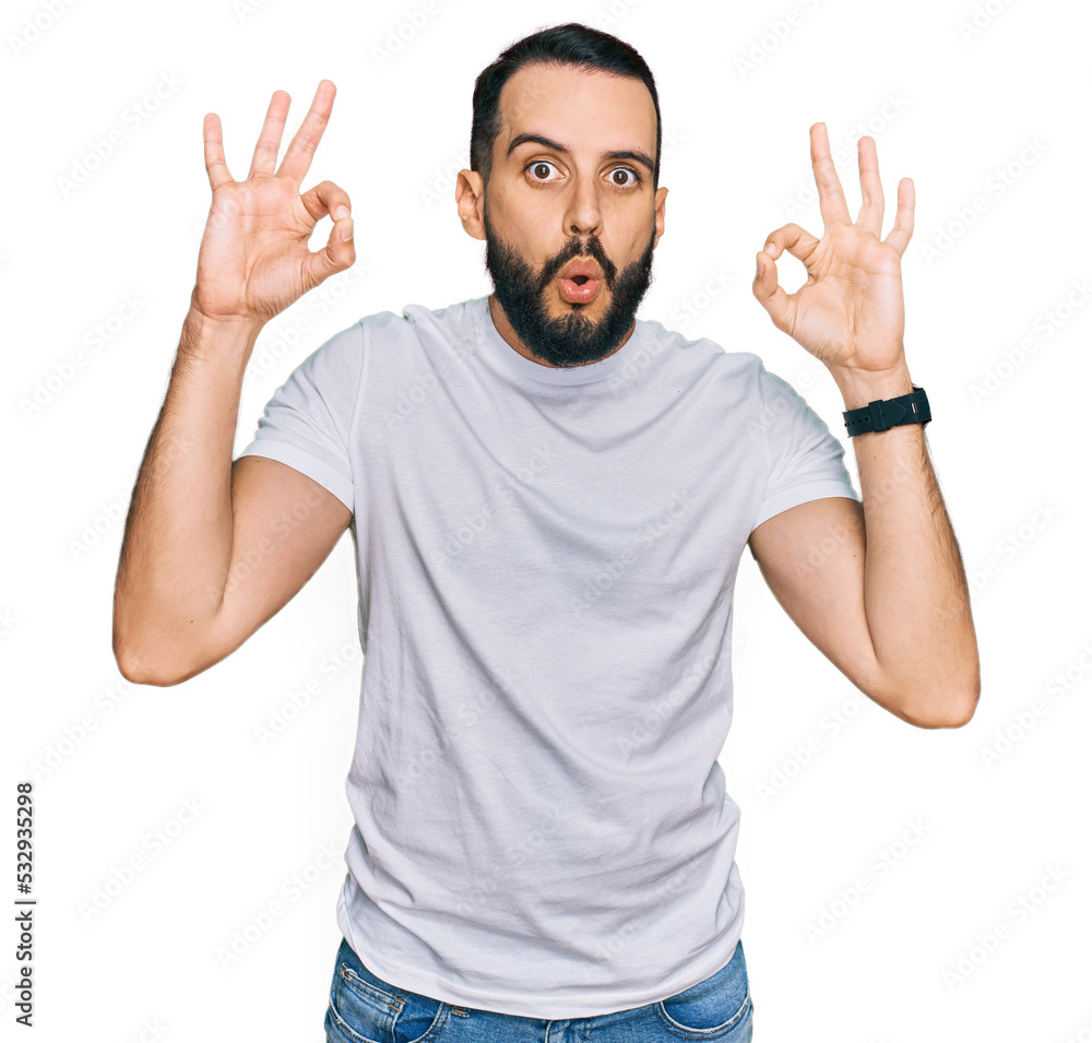 Young man with beard wearing casual white t shirt looking surprised and shocked doing ok approval symbol with fingers. crazy expression Young man with beard wearing casual white t shirt looking surprised and shocked doing ok approval symbol with fingers. crazy expression