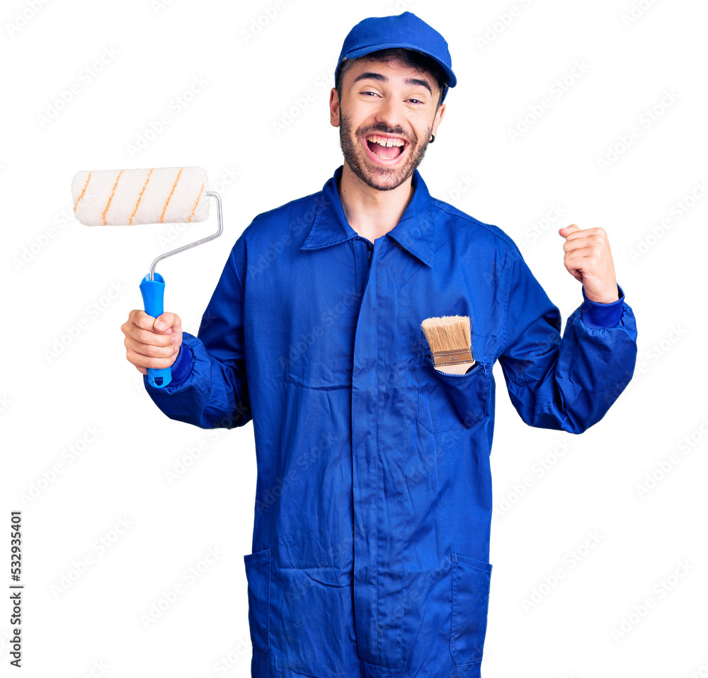 Young hispanic man wearing painter uniform holding roller screaming proud, celebrating victory and success very excited with raised arms