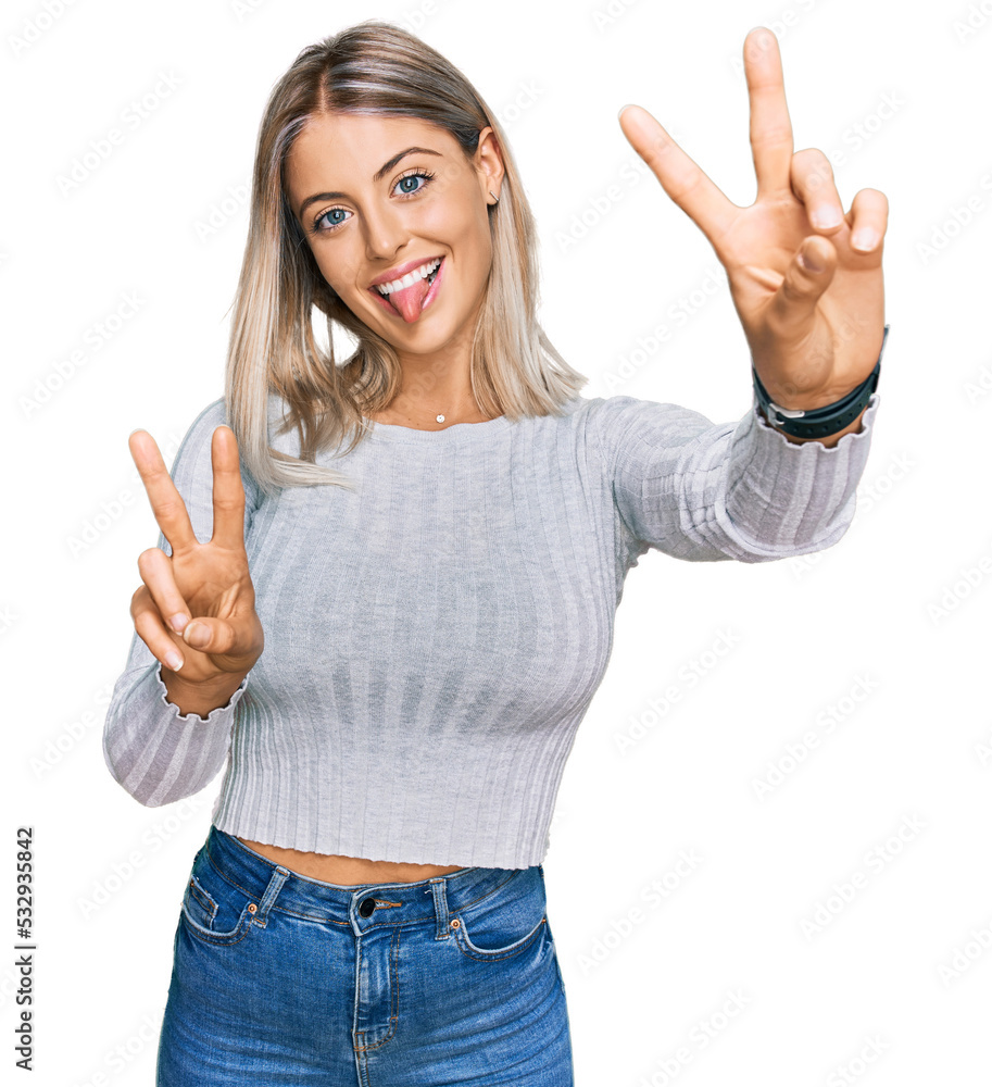 Beautiful blonde woman wearing casual clothes smiling with tongue out showing fingers of both hands doing victory sign. number two. Beautiful blonde woman wearing casual clothes smiling with tongue out showing fingers of both hands doing victory sign. number two.