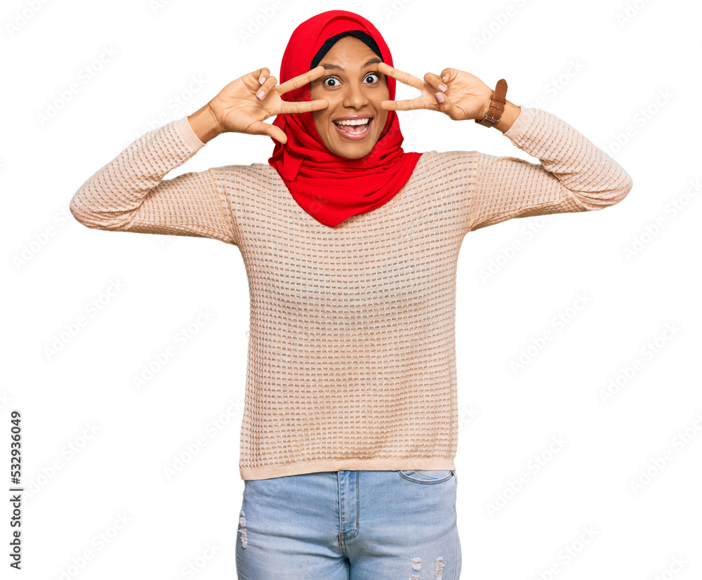 Naklejka premium Young african american woman wearing traditional islamic hijab scarf doing peace symbol with fingers over face, smiling cheerful showing victory