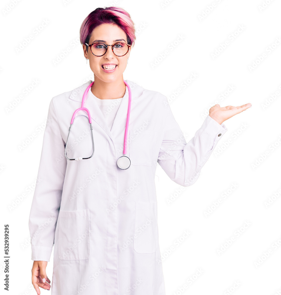 Young beautiful woman with pink hair wearing doctor uniform smiling cheerful presenting and pointing with palm of hand looking at the camera. Young beautiful woman with pink hair wearing doctor uniform smiling cheerful presenting and pointing with palm of hand looking at the camera.