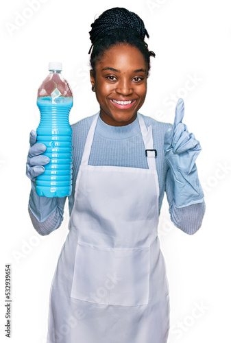 Young african american woman wearing apron holding detergent bottle surprised with an idea or question pointing finger with happy face, number one