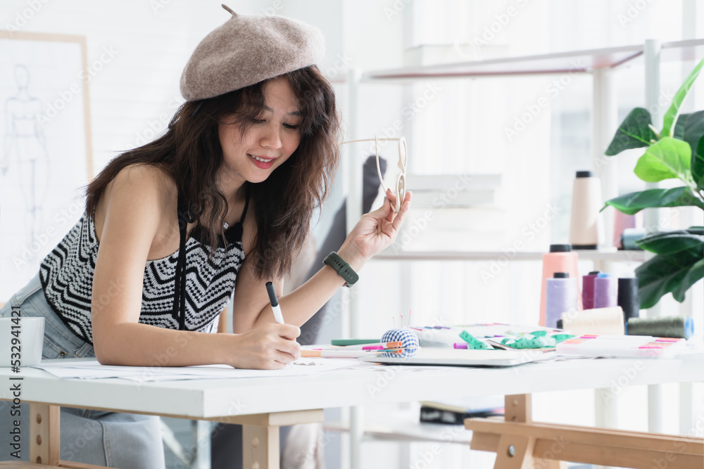 Asian attractive 30s woman fashion designer or dressmaker drawing sketch of new collection clothes on paper and smiling while working with tablet and tailoring equipments on desk at workplace studio Asian attractive 30s woman fashion designer or dressmaker drawing sketch of new collection clothes on paper and smiling while working with tablet and tailoring equipments on desk at workplace studio