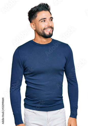 Fototapeta Young man with beard wearing casual blue winter sweater looking away to side with smile on face, natural expression