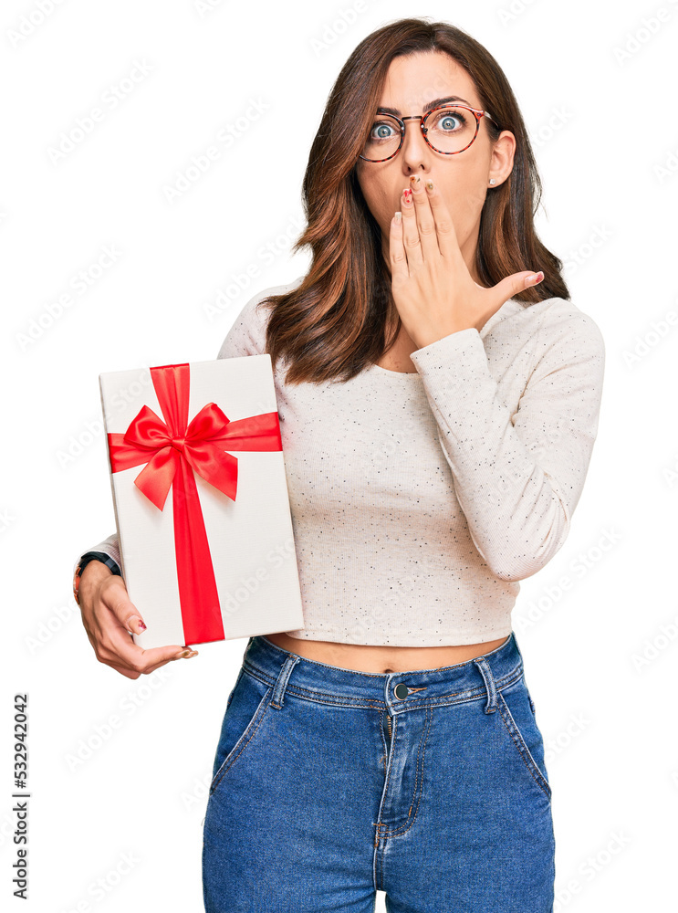 Young brunette woman holding gift covering mouth with hand, shocked and afraid for mistake. surprised expression Young brunette woman holding gift covering mouth with hand, shocked and afraid for mistake. surprised expression
