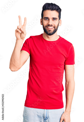 Young handsome man with beard wearing casual t-shirt showing and pointing up with fingers number two while smiling confident and happy.