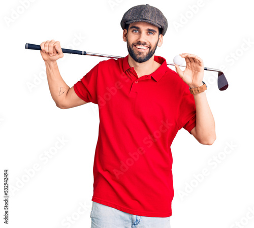 Young handsome man with beard playing golf holding club and ball looking positive and happy standing and smiling with a confident smile showing teeth