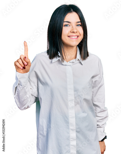 Young brunette woman with blue eyes wearing oversize white shirt showing and pointing up with finger number one while smiling confident and happy.