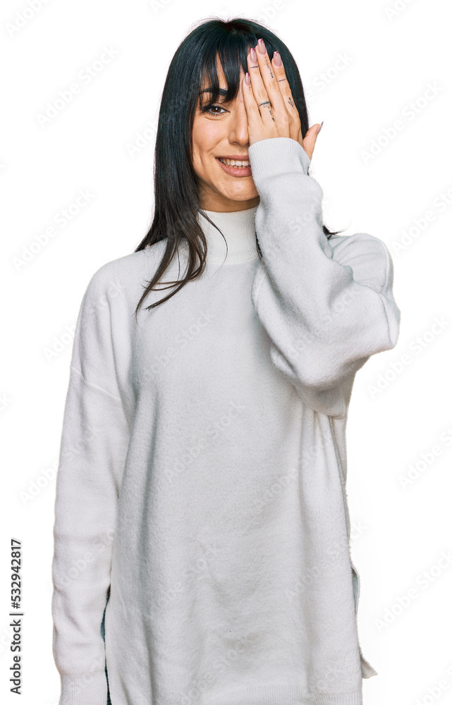 Young brunette woman with bangs wearing casual turtleneck sweater covering one eye with hand, confident smile on face and surprise emotion. Young brunette woman with bangs wearing casual turtleneck sweater covering one eye with hand, confident smile on face and surprise emotion.