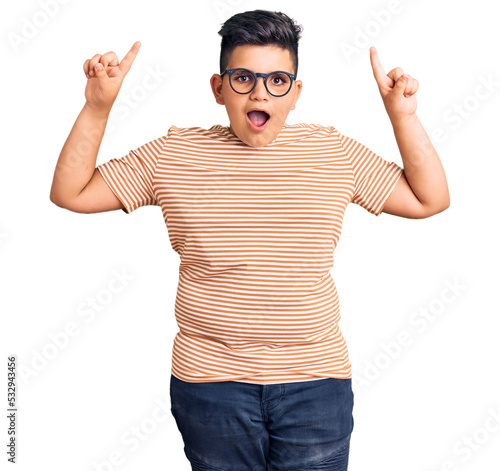 Little boy kid wearing casual clothes and glasses smiling amazed and surprised and pointing up with fingers and raised arms.