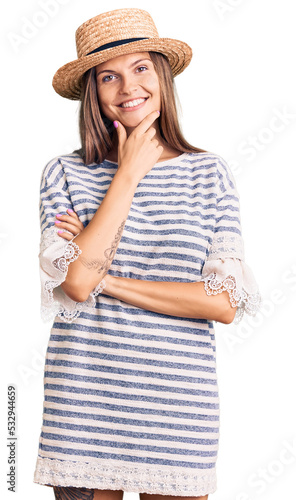 Beautiful caucasian woman wearing summer hat looking confident at the camera smiling with crossed arms and hand raised on chin. thinking positive.