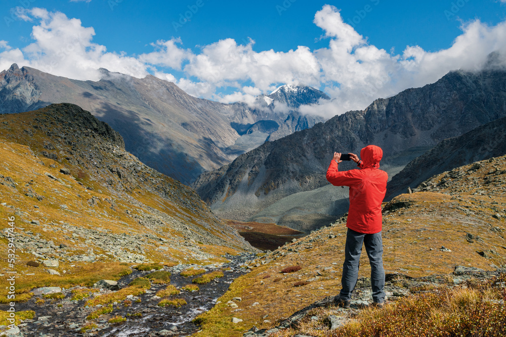 A man in a red jacket takes pictures of a mountain valley with his smartphone.