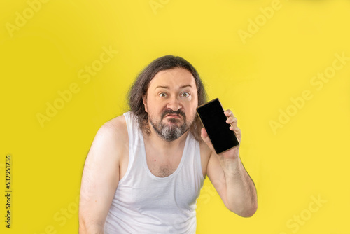 A disgruntled unshaven man in a white undershirt demonstrates a blank smartphone screen. An unemployed, unkemptly dressed adult from Eastern Europe.