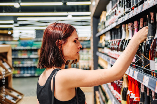 Young woman taking bottle of wine from shelf at supermarket
