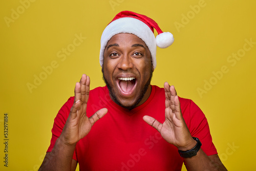 Shocked young African-American Santa Claus in a red T-shirt and a Christmas hat, shouting with a hand gesture at his mouth, isolated on a yellow background. Concept Happy New Year, Merry Christmas © Aleksandr