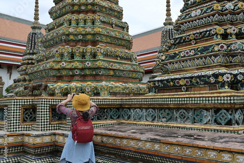 A female traveler wearing a yellow hat with a red backpack and jeans is using a mobile phone. Take photos of the magnificent pagodas in famous temples: Wat Pho or the Reclining Buddha Temple in Bangko photo