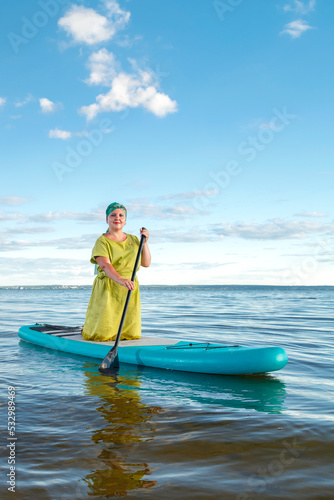 A woman in a dress and a headdress standing on a SUP board with an oar floats on the water against the blue sky.