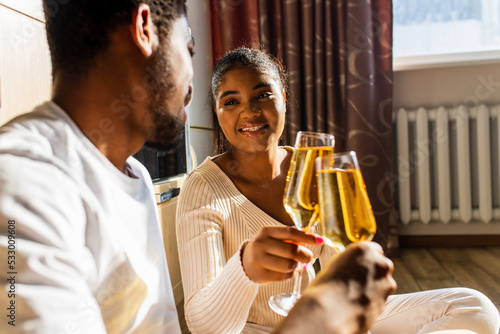 Laughing young afro ethnic couple dating with champagne at home while sitting at the warm floor