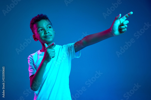 Studio shot of emotional little boy, kid expressing different emotions isolated over blue background in neon light. Education, childhood, modern lifestyle © master1305