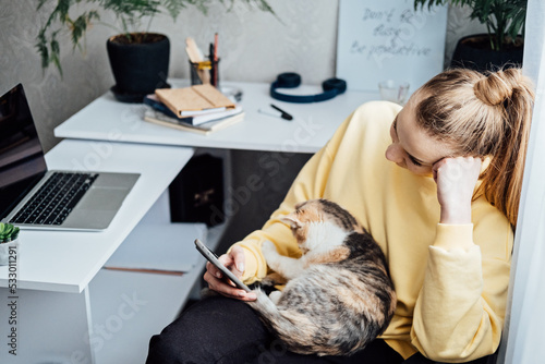 Woman freelance or procrastinate at workplace at home office. Self-employed businesswoman with cat distracted from work on laptop scrolling social media on smartphone. photo