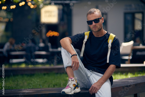 Young handsome bearded man in stylish black clothes looking through sunglasses, with a fashionable hairstyle, with backpack relaxing on the street. A handsome modern guy. Street style.
