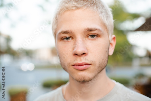 Handsome young man with blond short cut hairstyle smiling over city urban background outdoor. Look at camera. Close up portrait of 18-19 year old happy teenage boy.