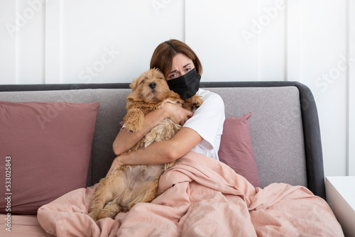 Young sad woman in medical mask lying in the bed and hugging her adorable pet dog. Perfect friendship despite allergy