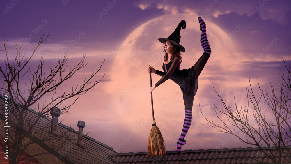 Foto Stock ballerina on pointe shoes in a black witch costume in a hat and  with a broom dances on the roof against the backdrop of the night sky and  the big