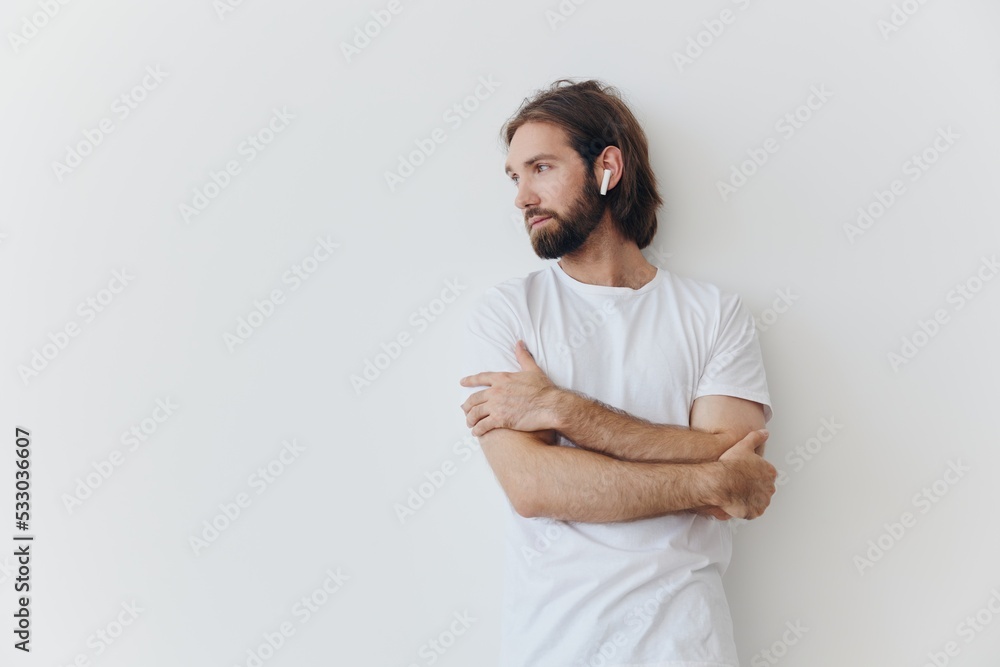 A man with a beard and long hair in a white T-shirt and blue jeans stands against a white wall, leaning against it and listening to music with wireless white headphones, staring thoughtfully A man with a beard and long hair in a white T-shirt and blue jeans stands against a white wall, leaning against it and listening to music with wireless white headphones, staring thoughtfully