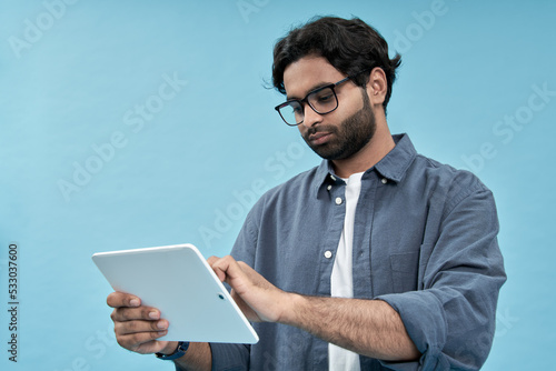 Young arab man student using digital tablet elearning standing isolated on blue background. Ethnic indian guy holding pad computer wearing glasses watching virtual class webinar, browsing internet.