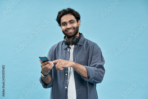 Happy young arab man using cell mobile phone isolated on blue background. Smiling ethnic guy holding smartphone playing game in app, paying online, betting, buying in e commerce shop on cellphone.