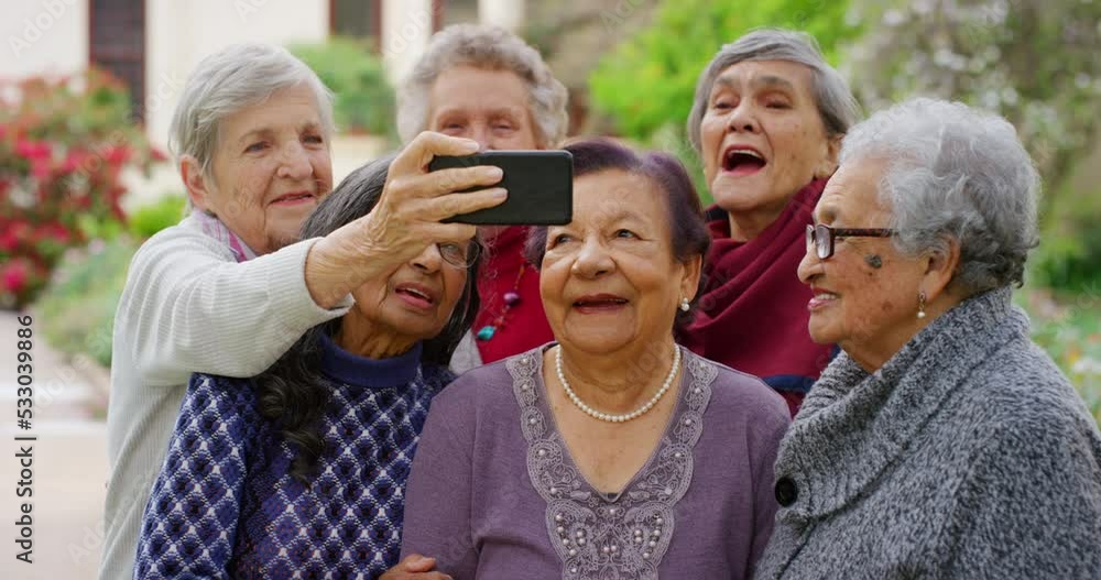 Group of elderly friends taking a selfie with a phone while standing in the retirement house garden. Happy senior women on a video call together on a smartphone in nature in the old age home park. Group of elderly friends taking a selfie with a phone while standing in the retirement house garden. Happy senior women on a video call together on a smartphone in nature in the old age home park.