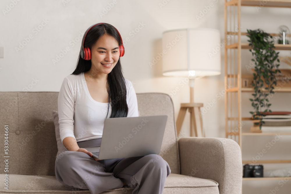 Smiling young asian woman enjoying free time while relaxing with her laptop in living room.. Smiling young asian woman enjoying free time while relaxing with her laptop in living room..