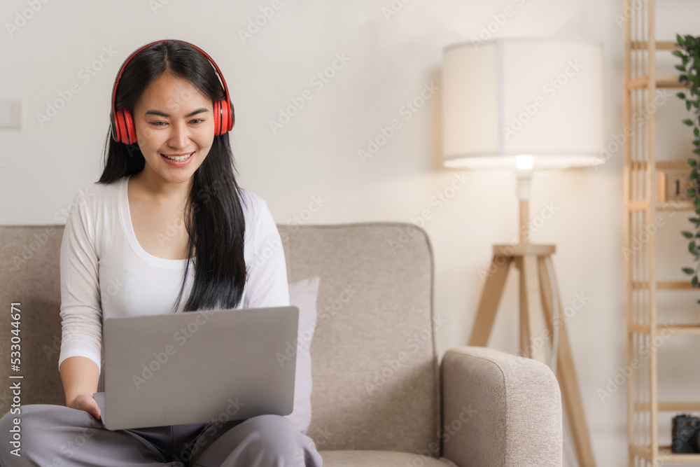 Smiling young asian woman enjoying free time while relaxing with her laptop in living room.. Smiling young asian woman enjoying free time while relaxing with her laptop in living room..