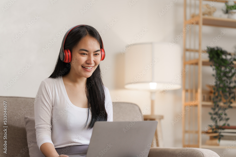 Smiling young asian woman enjoying free time while relaxing with her laptop in living room.. Smiling young asian woman enjoying free time while relaxing with her laptop in living room..