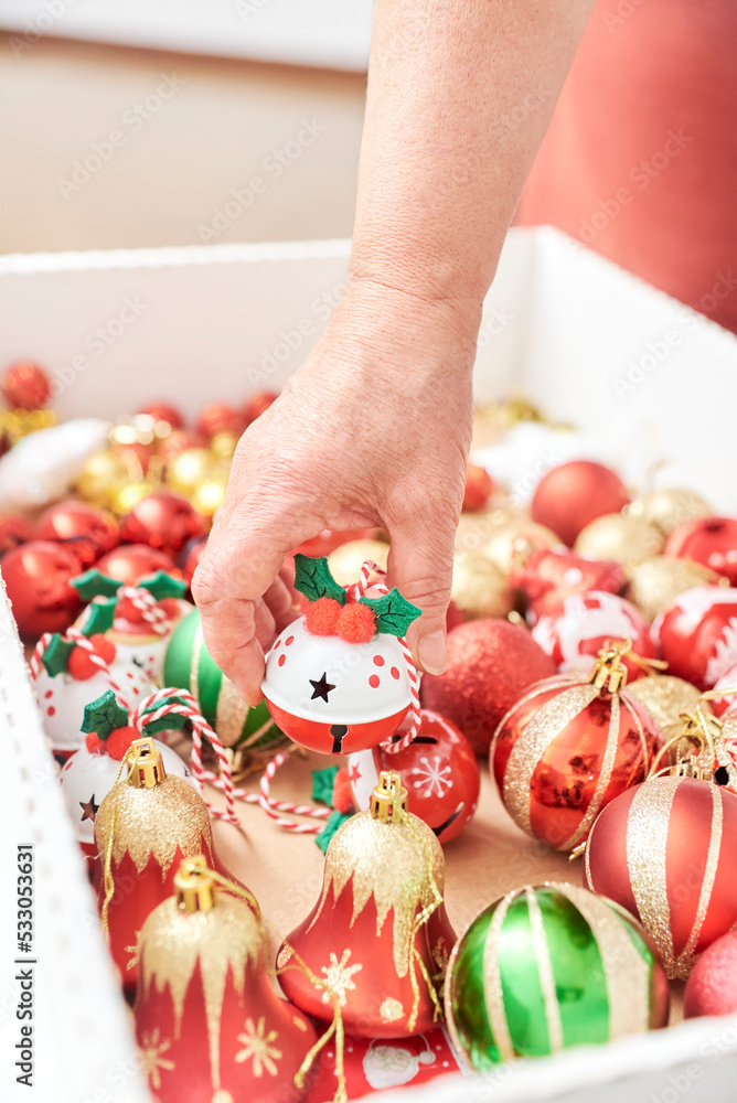 Female hand grabbing a ball from a box of mixed Christmas tree ornaments in the classic traditional colors of red, green, white and gold. Unrecognizable woman decorating at home.