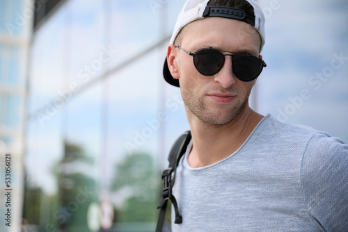 Handsome young man with stylish sunglasses and backpack near reflection surface outdoors  space for text