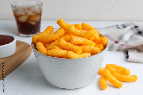 Crunchy cheesy corn snack in bowl, ketchup and refreshing drink on white wooden table, closeup