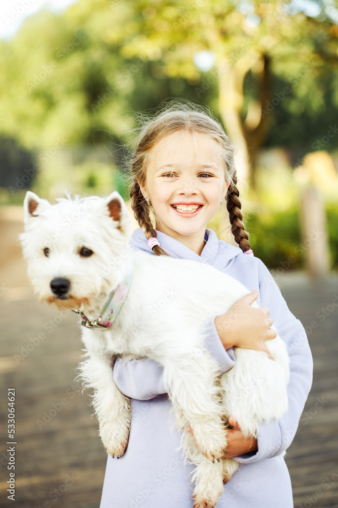 A girl with pigtails holds a small white dog in her arms and smiles happily. A girl with pigtails holds a small white dog in her arms and smiles happily.