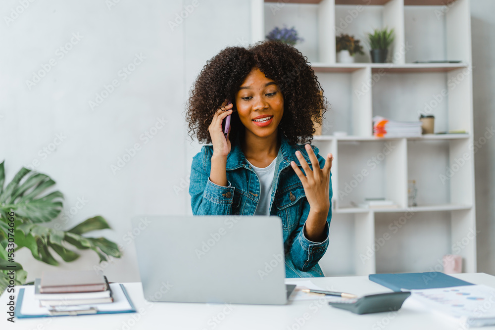 Young female freelancer talking on phone and using laptop at desk. Young female freelancer talking on phone and using laptop at desk.