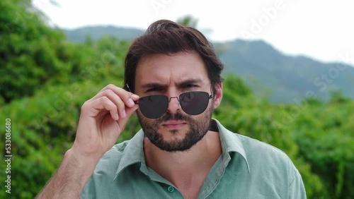 Attractive bearded dark-haired young Hispanic man looks at the camera through his lowered sunglasses, then removes them and covers his mouth, expressing astonishment and bewilderment at what he saw