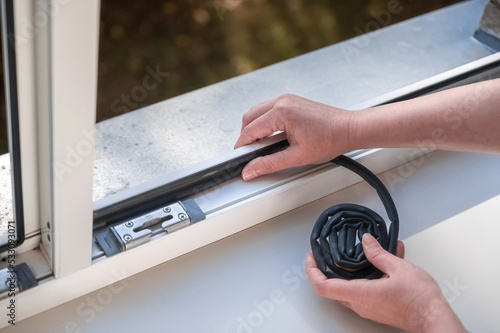 Preparing for the cold season. A woman sticks a dark rubber sealing tape on a window indoors.