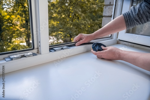 Preparing for the cold season. A woman sticks a dark rubber sealing tape on a window indoors.