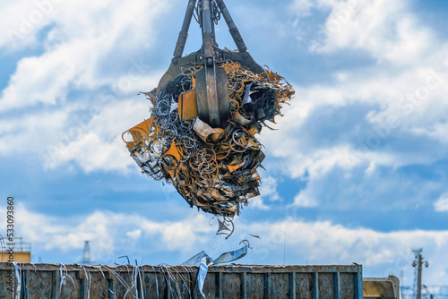 A grab crane loads iron, steel and scrap metal into a truck for recycling at an industrial waste collection center with small falling particles of metal chips and dust. Transportation of metal waste.