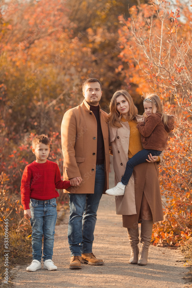 Happy family with two kids walking in the fall park. Full body portrait of a caucasian mother and father children in beautiful fashion outfits on sunny autumn day in forest. Family lifestyle concept.