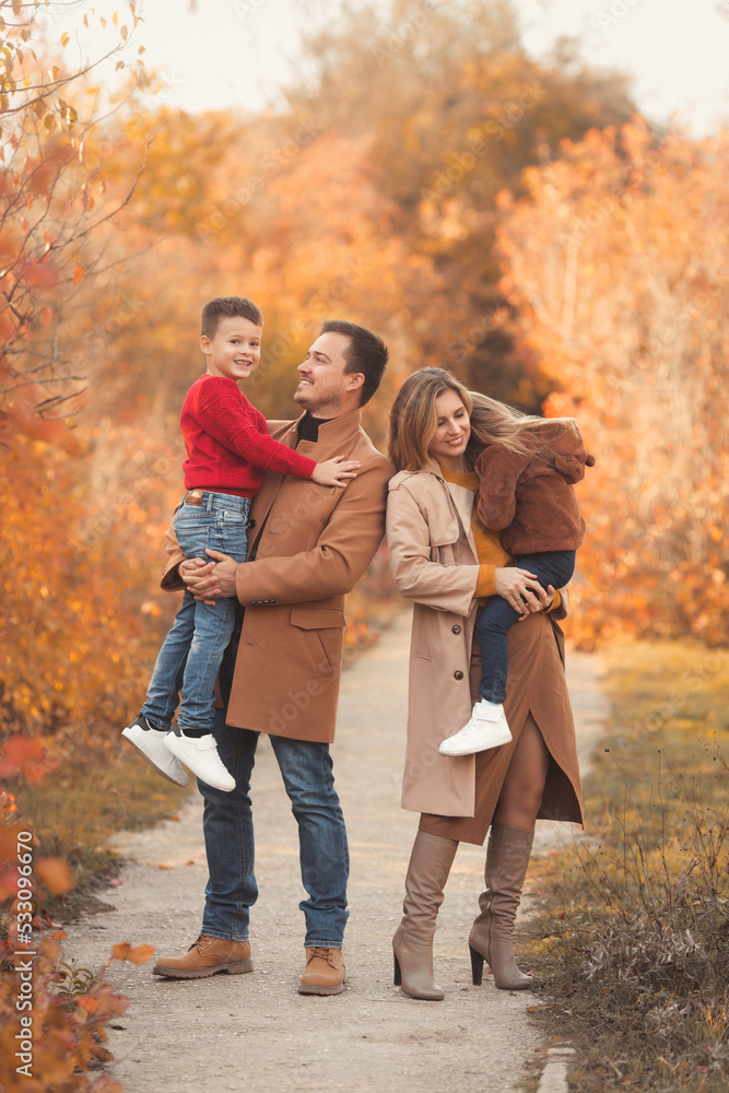 Happy family of four walking in the fall park. Portrait of a caucasian mother and father hugging their children in beautiful stylish outfits on a sunny autumn day in forest