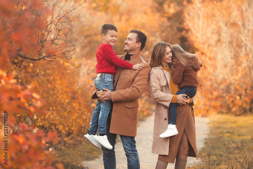 Smiling and happy family of four: mom and dad with two kids hugging together in fashion outfits, walking in fall forest
